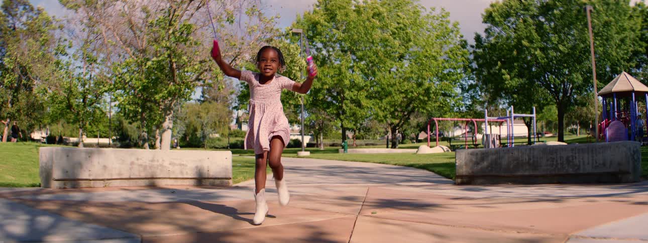 A joyful young girl in a dress and boots happily jumps rope in slow motion at a sunny park