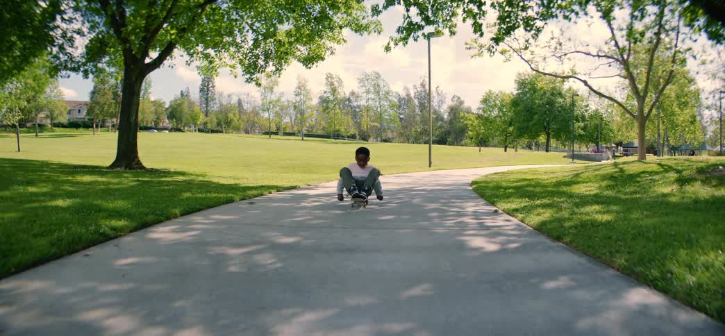 A young boy smiles as he skates towards the camera while sitting on his board on a path in a sunny green park