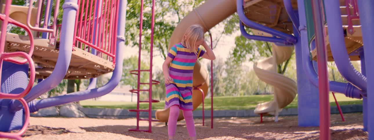 A young girl dressed in bright colors tries to hula hoop in slow motion at an outdoor playground but it falls down