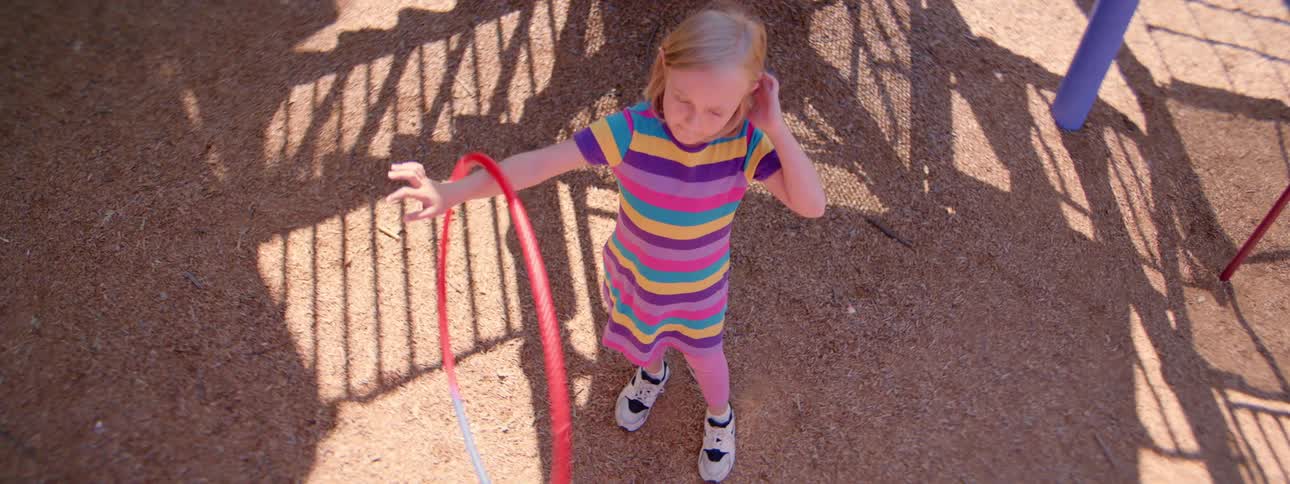 A young girl in a colorful striped dress spins a hula hoop on her arm in slow motion at a sunny playground, shot from a high angle