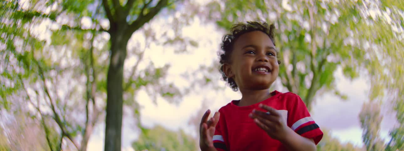 An adorable toddler boy in a red shirt laughs and smiles with excitement while playing in a park in slow motion