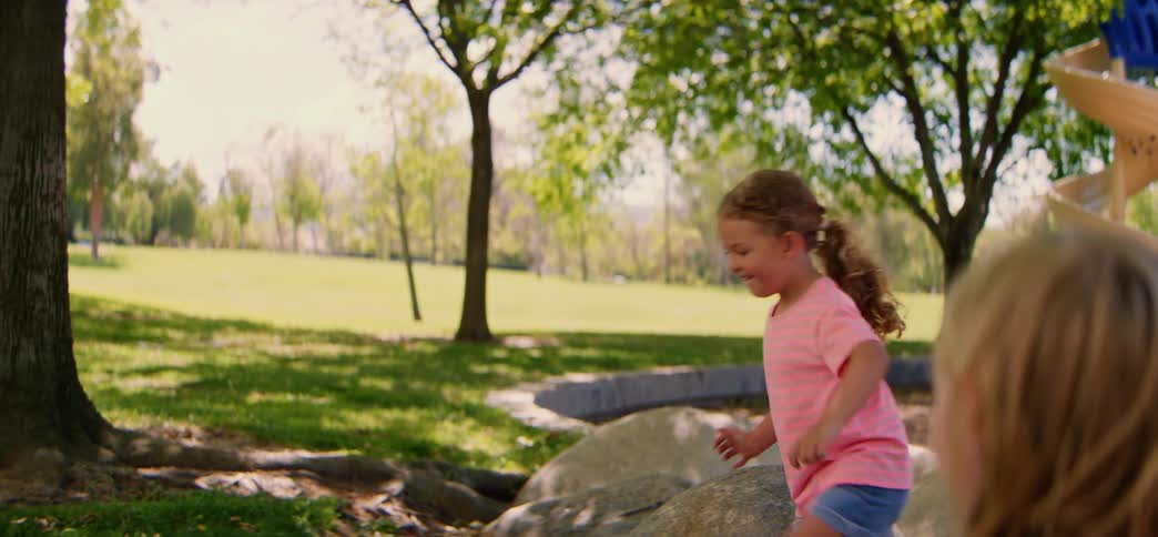 A cheerful young girl carefully balances and walks across a series of large rocks in a sunny park before her friend offers a helping hand