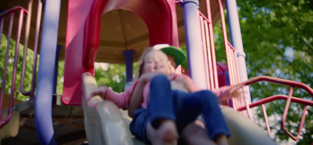 A happy father holds his little girl as they slide down a playground slide in slow motion