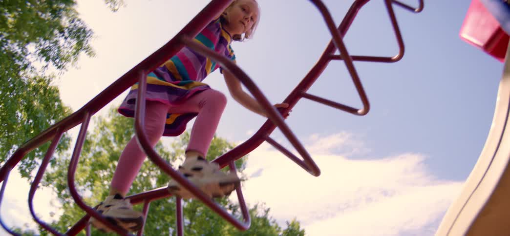 From a low angle, a little girl in a colorful dress joyfully climbs a red ladder on a sunny playground in slow motion