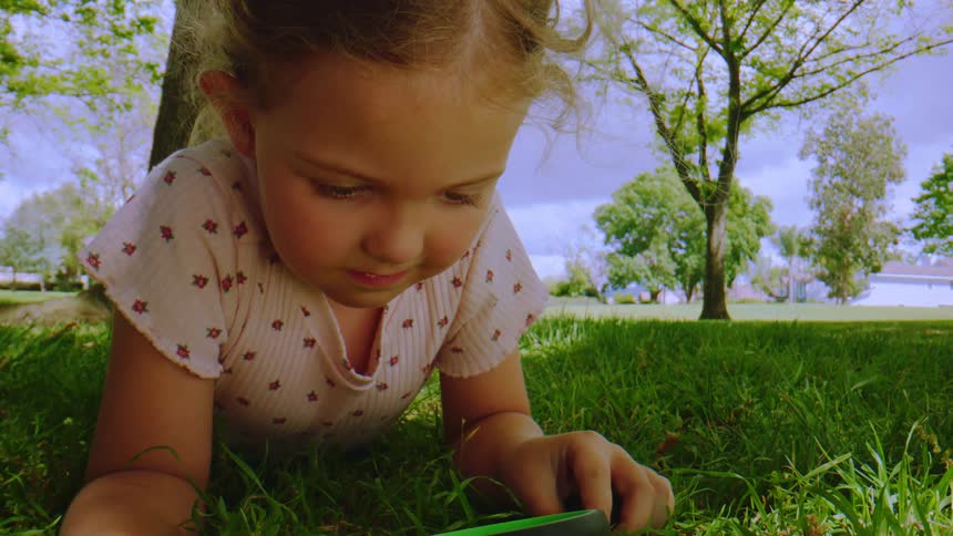 A curious little girl lies in the bright green grass at the park, intently studying something on the ground