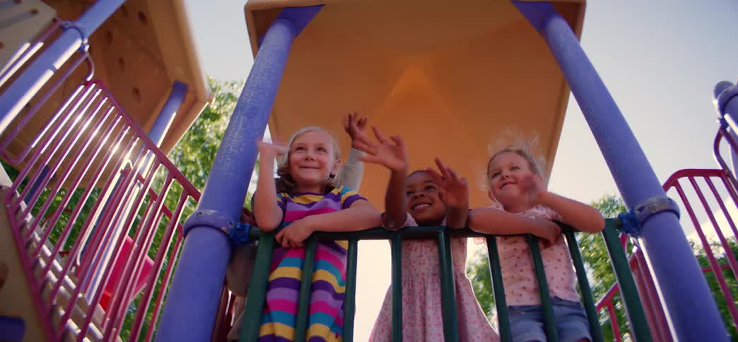 From a low angle, a happy and diverse group of young children wave down from a colorful playground structure