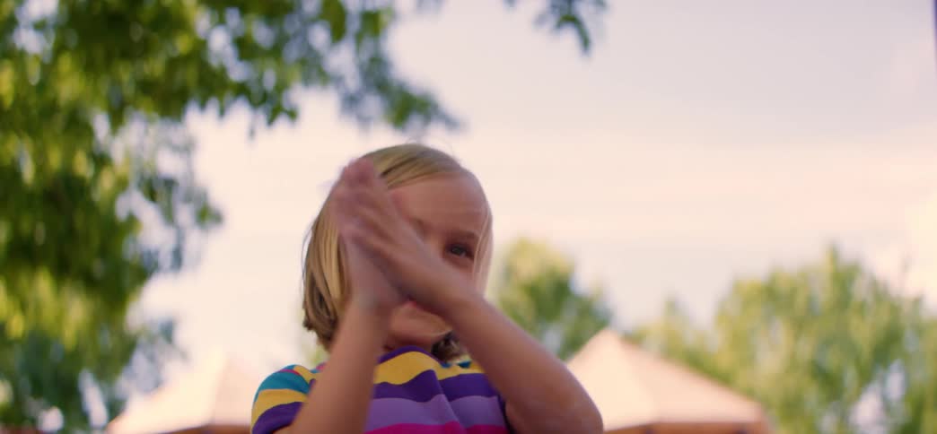A smiling young girl in a colorful striped shirt claps her hands while looking at the camera before turning and running away in slow motion