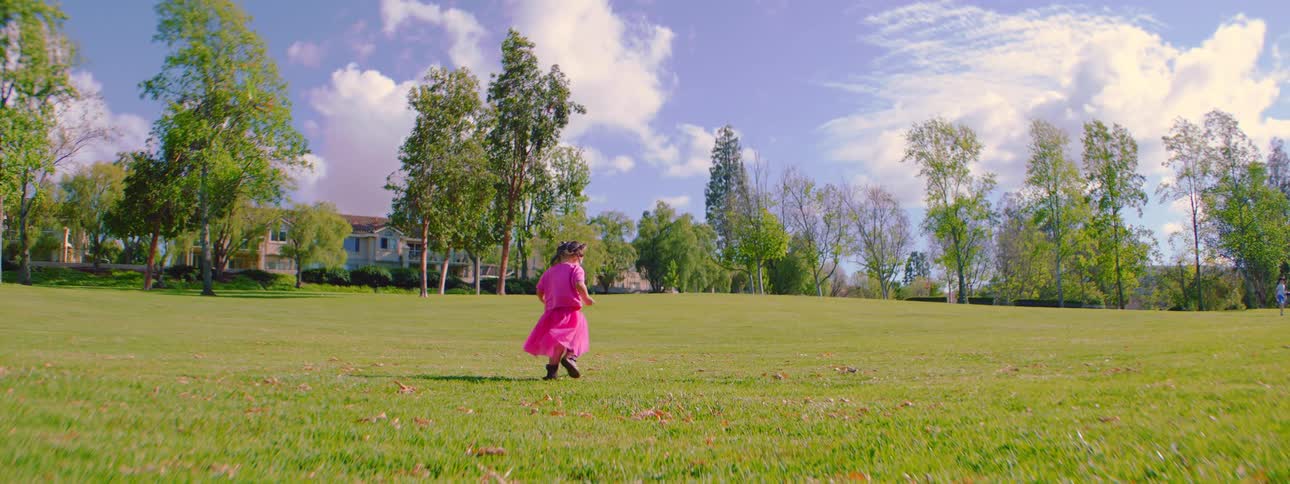 A little girl in a vibrant pink dress runs in slow motion across a vast, sunlit green park