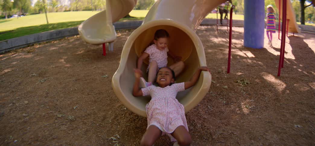 Three smiling young girls slide down a tunnel slide one after another at a sunny playground in slow motion