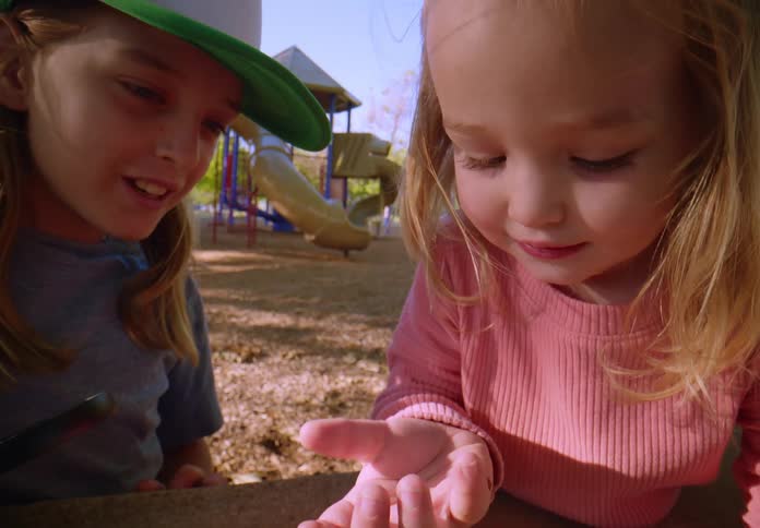 Two curious young sisters examine a small bug crawling on a hand at the playground on a sunny day