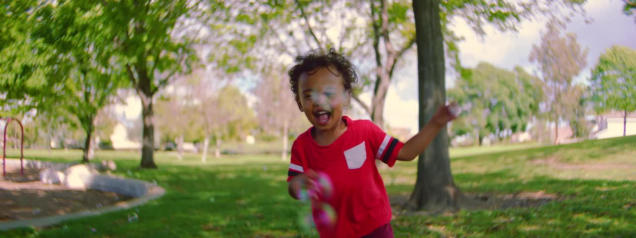 Slow motion shot of a joyful toddler with curly hair running through a sunny park and playing with soap bubbles