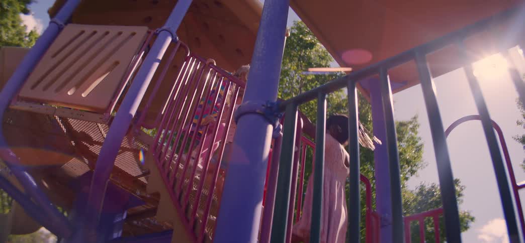 A dreamy low-angle shot shows two diverse little girls climbing up a colorful playground structure on a sun-drenched day