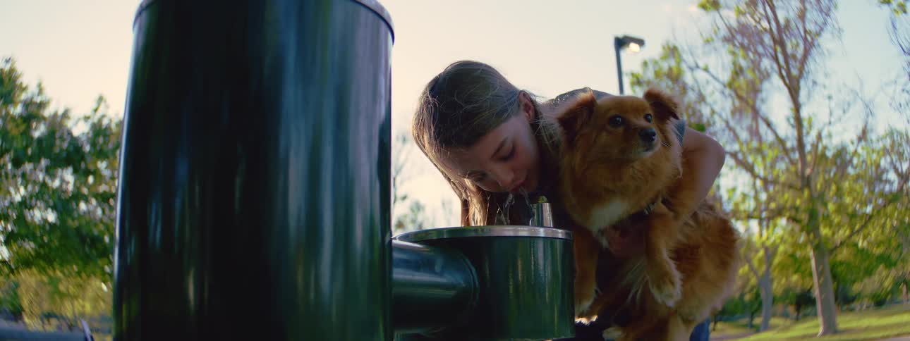 A young girl holds her fluffy brown dog while taking a refreshing drink from a water fountain in a sunlit park