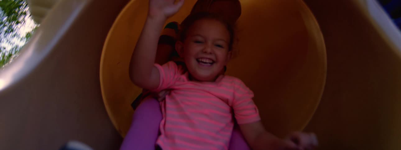 Two happy young girls slide down a yellow tube slide together at the playground in slow motion