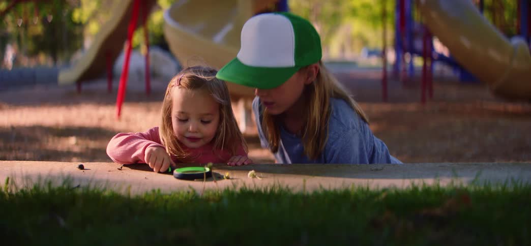 A young girl and her older sister discover the wonders of nature together using a magnifying glass at a park playground
