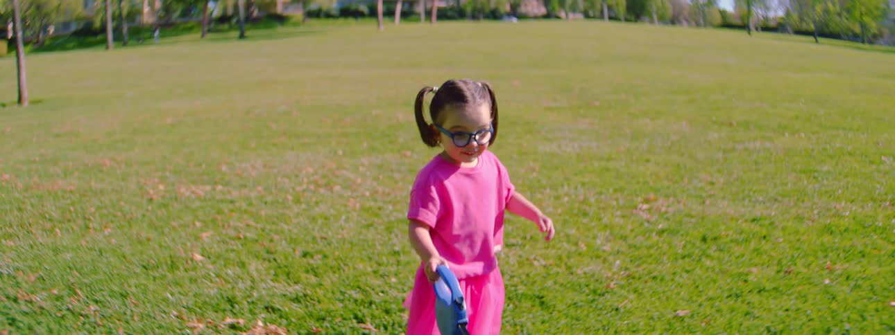 A cute little girl in a pink dress and glasses walks her small fluffy dog across a green grassy field on a sunny day in slow motion