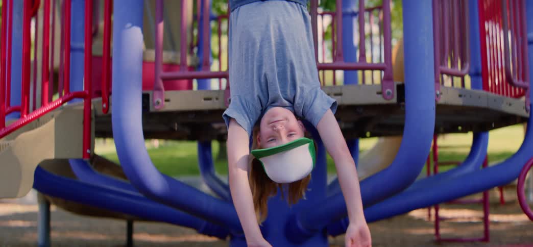 A happy young boy in a trucker hat smiles at the camera while hanging upside down on playground equipment