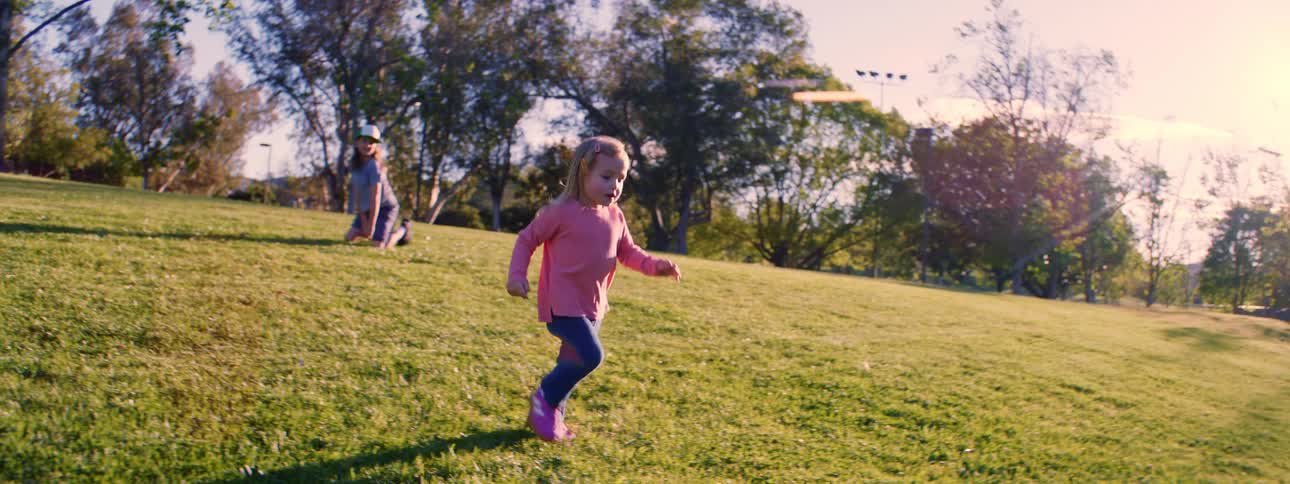 A joyful little girl with blonde hair runs across a sunny, grassy hill in a park in super slow motion