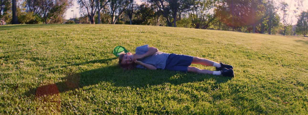 A carefree young boy with long hair rolls around and relaxes on a sun-drenched grassy hill in a park