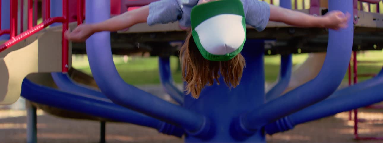 Close up of a happy young boy in a trucker hat hanging upside down at a colorful playground in slow motion