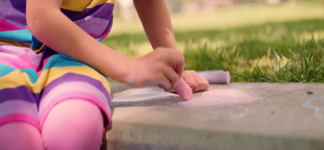 A close-up shot of a young girl's hands coloring with pink chalk on the sidewalk next to a green lawn