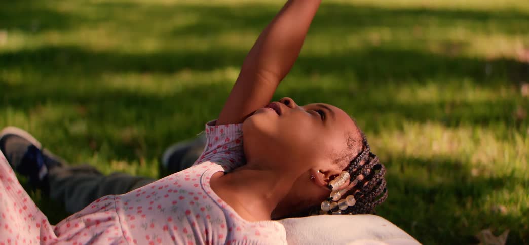 A heartwarming close-up of a young girl with braided hair laughing and playing while lying in the grass on a sunny day