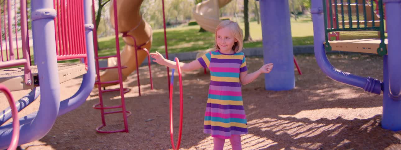 Slow motion shot of a smiling little girl in a vibrant striped dress skillfully spinning a hula hoop on her arm at the park