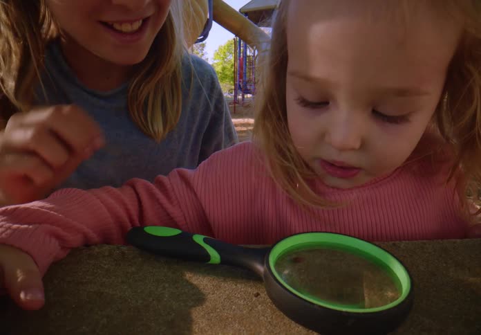 Two curious young sisters use a magnifying glass to get a closer look at ladybugs at the park
