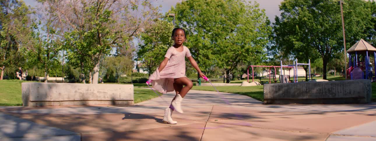 A joyful young girl in a dress and boots skillfully jumps rope in slow motion at a sunny park