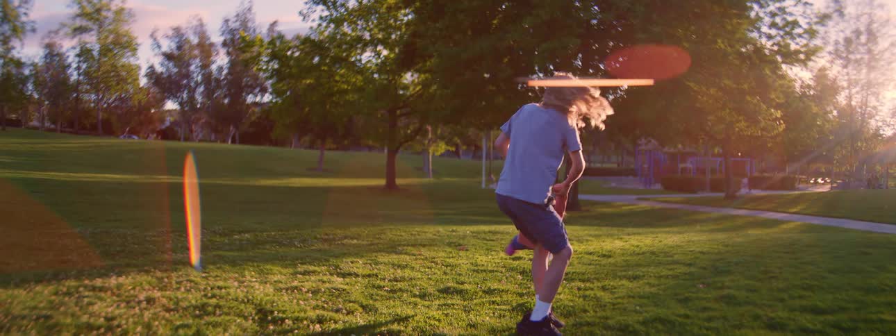 A long-haired brother playfully swings his younger sister around in a sunlit park in slow motion