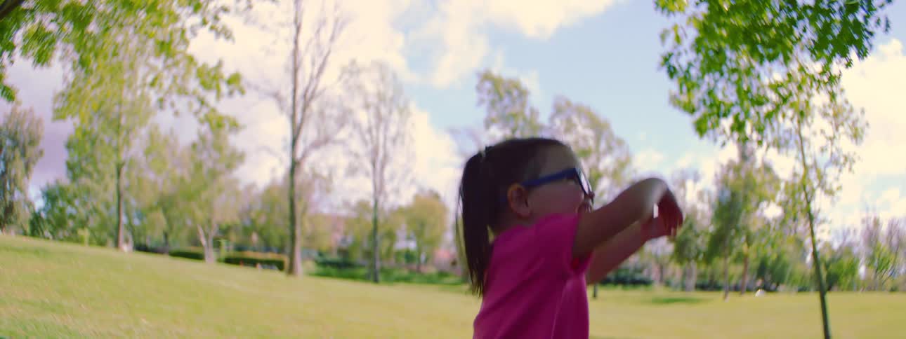 In super slow motion, a delighted little girl with pigtails and glasses runs and jumps through a sunny park, chasing after bubbles