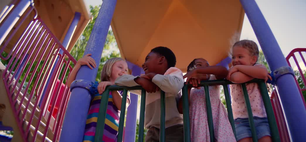 A diverse group of four happy children laugh and smile together while playing on a jungle gym at a park