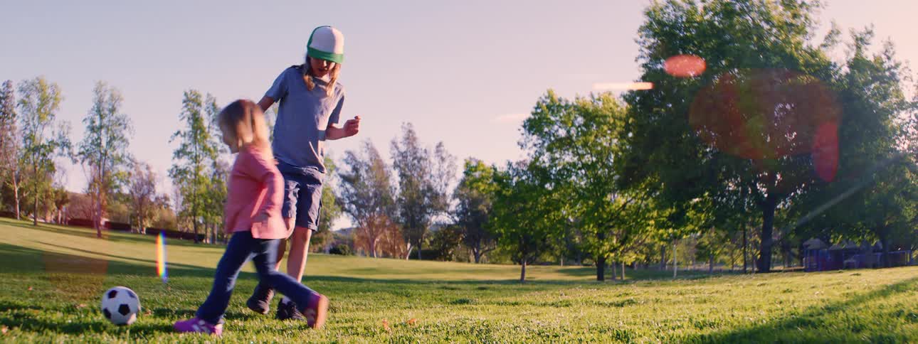 A young boy and girl playfully chase a soccer ball across a grassy field with a warm lens flare in slow motion