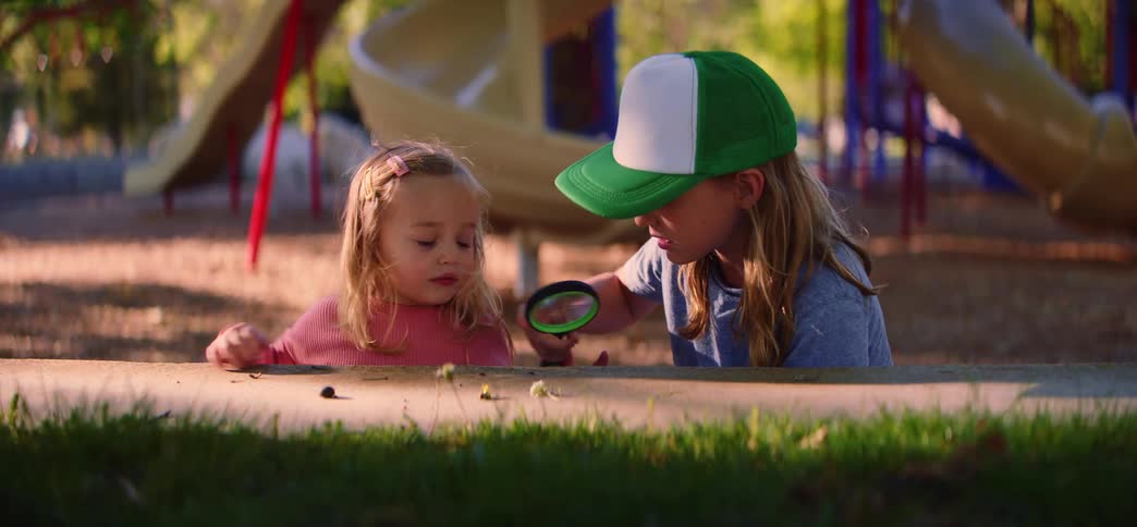Two young sisters curiously examine a bug together with a magnifying glass at the park playground