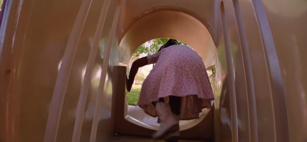 A joyful young girl with beaded braids playfully crawls through a playground tunnel and turns to smile at the camera in slow motion