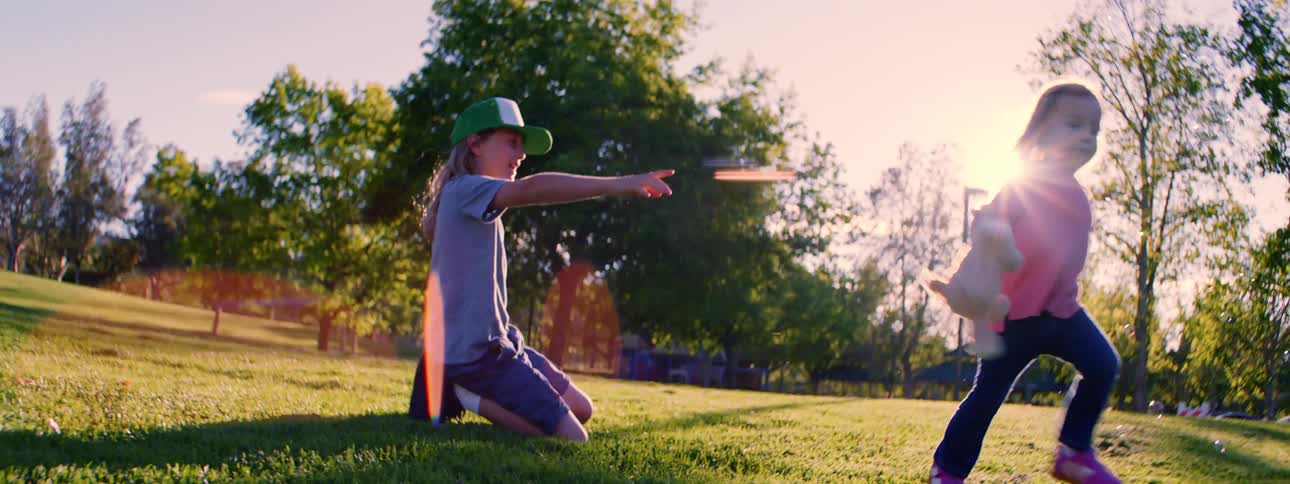 A young boy throws a frisbee in a sunny park while his little sister runs by with a stuffed animal, filmed in slow motion