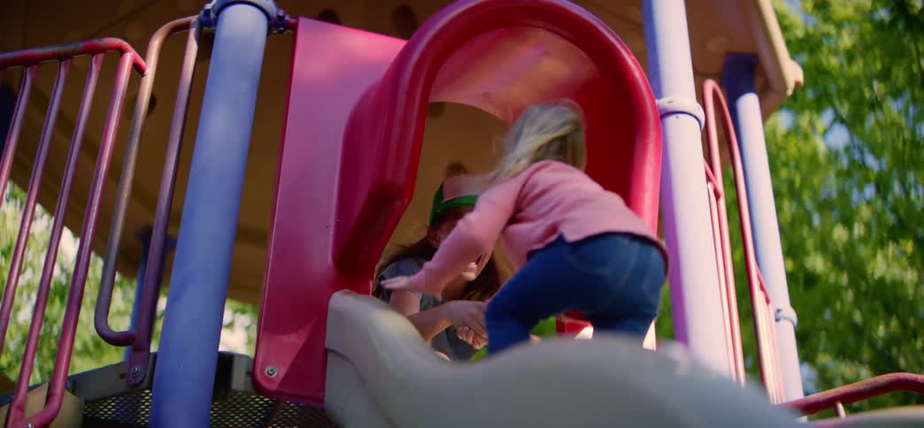 Two young girls laugh as they play together on a colorful slide at the park in slow motion