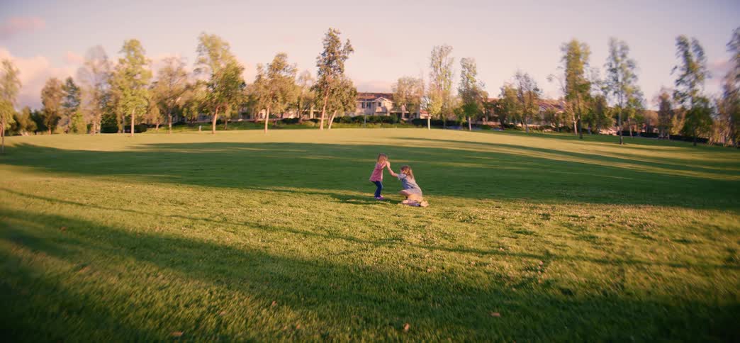 In slow motion, a brother and his toddler sister run and play together in a wide, grassy park during a beautiful golden hour