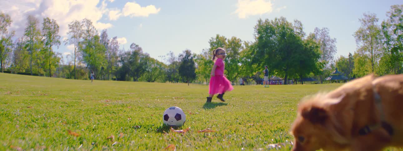 In slow motion, a little girl in a bright pink outfit runs across a sunny, green field toward a soccer ball, followed by her curious dog