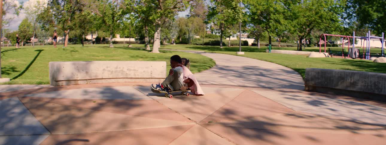 A young girl hugs her brother as they ride a skateboard together down a path in a sunny park