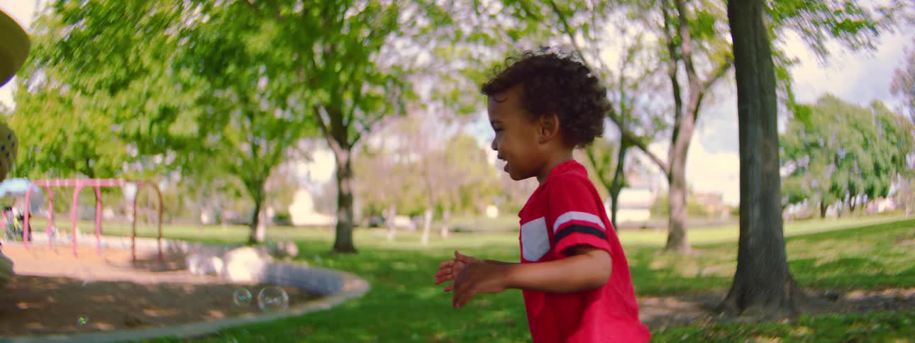 A joyful toddler with curly hair laughs and runs through a sunny park in slow motion, playing with soap bubbles as a little girl watches in the background