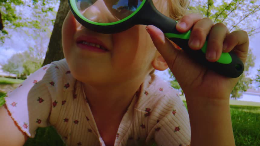 Slow motion close up of a curious little girl looking at the camera through a magnifying glass in a park