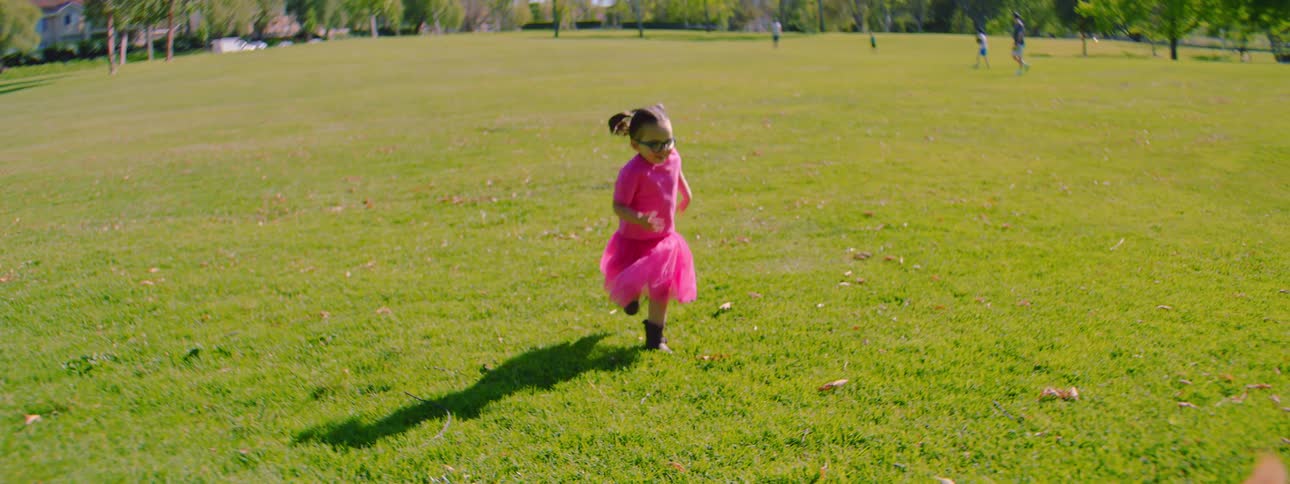 A little girl in a bright pink tutu runs in slow motion across a sunny green park and plays with her small fluffy dog