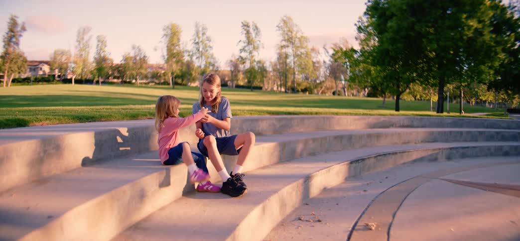 An older brother shares his snack with his younger sister as they sit together on concrete amphitheater steps in a park during a sunny afternoon