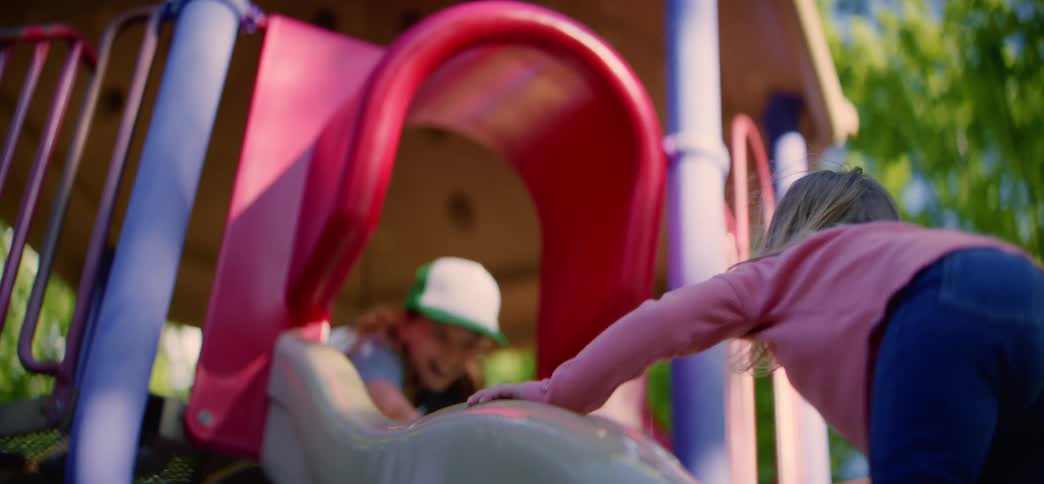 A joyful little girl climbs up a playground slide where her smiling friend is waiting, and then she slides down happily
