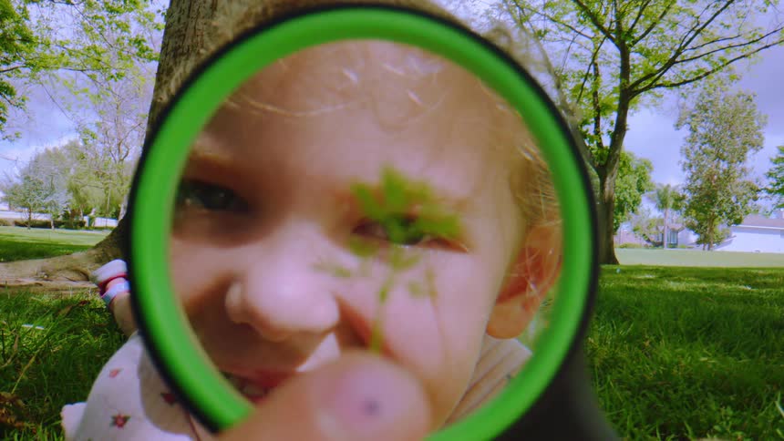A smiling young girl's face is seen through a magnifying glass as she curiously examines a small green plant while lying in a grassy park