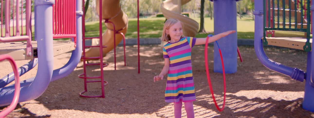 A smiling young girl in a colorful striped dress spins a hula hoop on her arm at the playground in slow motion