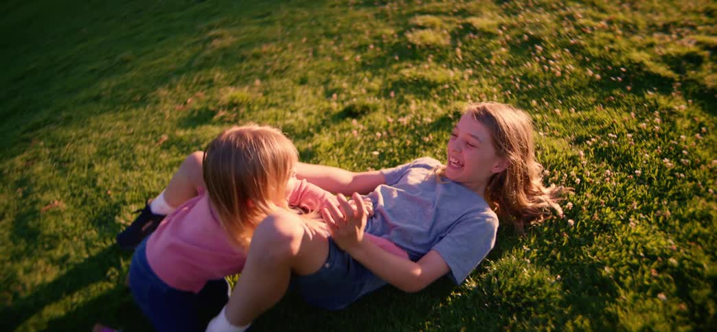 A happy little girl runs in slow motion across a green park and playfully tackles her older sister, laughing as they fall onto the grass together