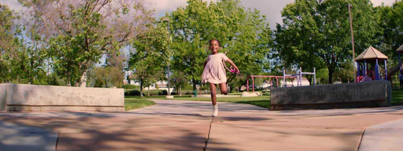 A joyful young girl wearing a pink dress happily jumps rope in slow motion towards the camera in a park on a sunny day