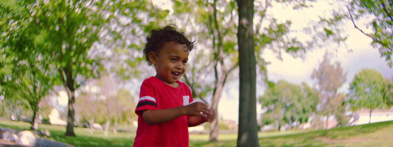An adorable toddler in a red shirt giggles with delight while chasing and playing with soap bubbles in a park in slow motion